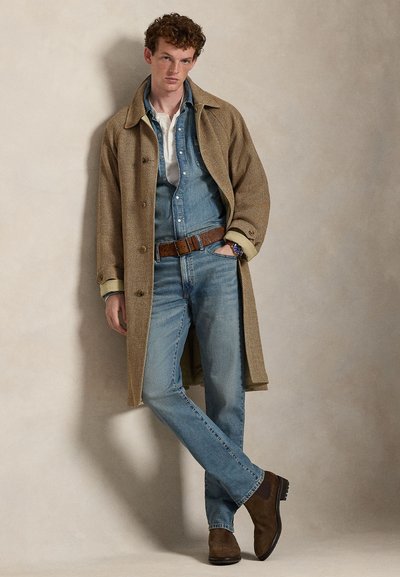 Jonge man leunt tegen de muur, draagt een lange bruine jas, denim overhemd en spijkerbroek, bruine riem en bruine laarzen, met één hand in de zak.