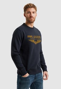 Mladič modri pulover iz mehkega materiala, s krožnim ovratnikom, ribastimi manšeti in z zlatim grafičnim tiskom z krili ter besedilom spredaj.