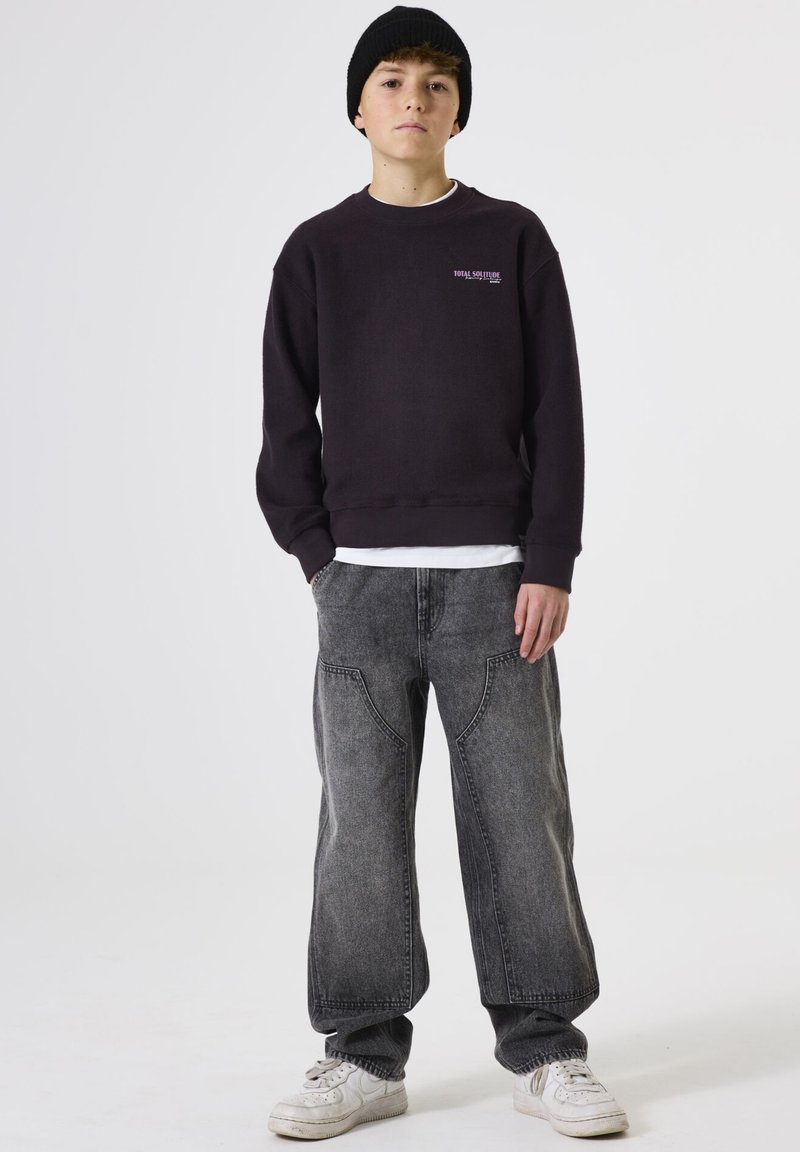 Sudadera de color púrpura oscuro con cuello redondo, camiseta blanca debajo, jeans de pierna ancha grises y zapatillas blancas. Gorro de lana negro.