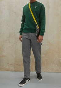 Grön Nike-tröja, randiga svart-vita byxor, svarta sneakers med vit accent och en liten svart väska med en gul rem.