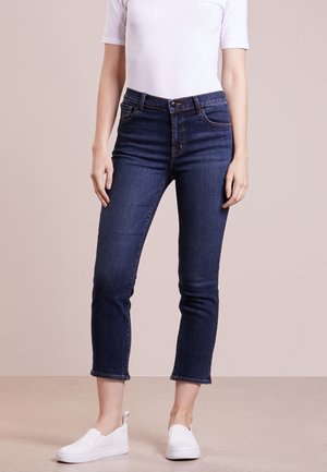 Personne portant un jean court bleu foncé, un haut blanc à manches courtes et des chaussures blanches sans lacets, debout devant un fond uni.