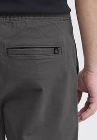 Graue Baumwollhose mit einer Gesäßtasche mit horizontaler Öffnung und einem kleinen runden Logosymbol neben der Tasche.