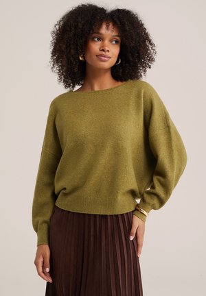 Femme aux cheveux bouclés, portant des boucles d'oreilles créoles dorées, un pull vert olive et une jupe plissée marron foncé sur un fond uni.