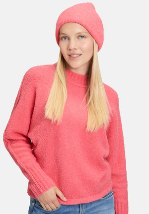Vrouw met lang blond haar, gekleed in een roze gebreide muts en een gestructureerde roze trui, met één hand in de zak van haar blauwe spijkerbroek tegen een witte achtergrond.