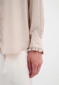 Blusa beige con una texture liscia, caratterizzata da maniche lunghe e dettagli di polsino arricciato. L'orlo è dritto e termina sotto la vita.