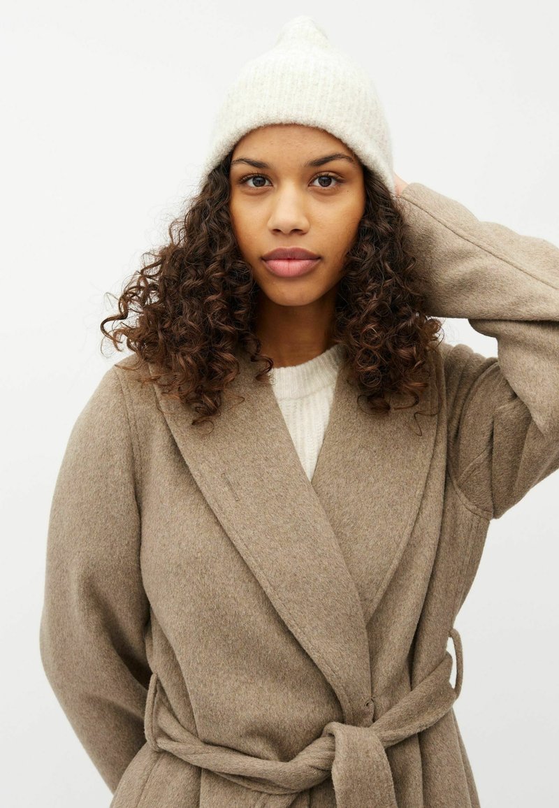 Cappotto di lana beige con design avvolgente, tessuto testurizzato, ampi rever e cintura. Il modello indossa un berretto di lana chiaro.