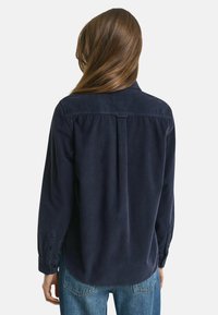Donna con lunghi capelli castani che indossa una camicia di velluto a coste blu scuro e jeans blu, vista di spalle su uno sfondo semplice.