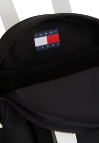 Εσωτερικό μαύρου υφασμάτινου σακιδίου με διχτυωτή θήκη. Διαθέτει ετικέτα σε μπλε, λευκό και κόκκινο με το κείμενο "TOMMY JEANS". Κλείνει με φερμουάρ.