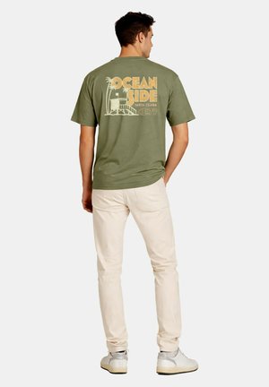 Hombre con camiseta verde oliva con gráfico "Oceanside Santa Clara" y pantalones beige, de pie de espaldas sobre un fondo liso.