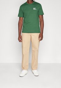 Zelené bavlněné tričko s bílým nápisem "UMÍTE MLUVIT LACOSTE?" na hrudi, spárované se světlými béžovými kalhotami a bílými teniskami.