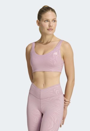 Donna che indossa un top sportivo viola chiaro e leggings coordinati a vita alta con motivo sottile, in piedi con le mani dietro la schiena e sguardo rivolto a destra.