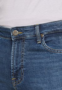 Pantalones vaqueros Lee de mezclilla azul con costuras amarillas y un botón metálico con la marca en la cintura. Textura suave, diseño clásico de cinco bolsillos.