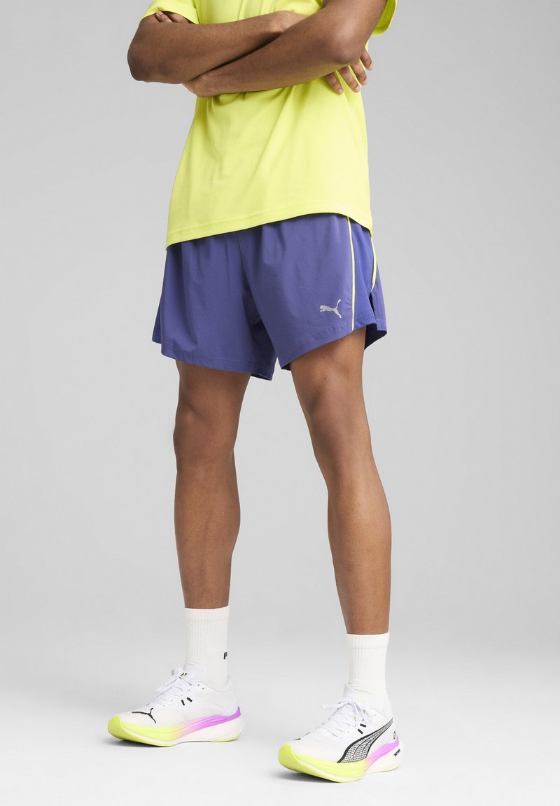 Lila Sportshorts mit gelben Akzenten und grauem Logo; kombiniert mit weißen Schuhen, die einen pinken und grünen Farbverlauf aufweisen. Helles gelbes T-Shirt.