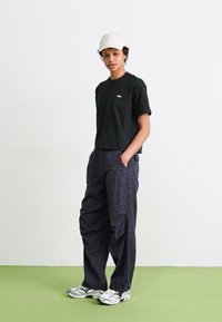 Svart t-skjorte med hvitt logo, marineblå rutete bukser, grå sneakers og en lys caps. Modellen står på grønt underlag mot en hvit bakgrunn.