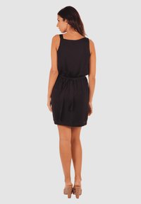 Vestido negro sin mangas con cintura ajustada y detalle de cinturón. Tela suave con un diseño simple, hasta la rodilla, combinado con tacones de color claro.