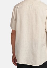 Camisa de manga corta beige claro con un ajuste relajado. Presenta una tela suave y texturizada y un dobladillo redondeado, mostrando una vista trasera lisa.