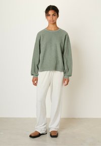 Maglione verde con una texture, maniche lunghe e scollatura rotonda, abbinato a pantaloni grigi chiari a vestibilità comoda e scarpe marroni senza allacciatura.