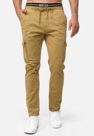 Man draagt beige cargobroek met trekkoord in de taille en witte sneakers, handen ontspannen langs het lichaam tegen een lichte achtergrond.
