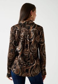 Una donna con lunghi capelli scuri indossa una camicia a maniche lunghe marrone e nera con motivo paisley e jeans blu, girata di spalle rispetto alla fotocamera.