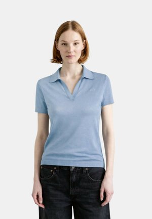 Giovane donna con capelli rossi corti che indossa una polo lavorata a maglia azzurro chiaro a maniche corte e jeans neri, in piedi davanti a uno sfondo semplice.