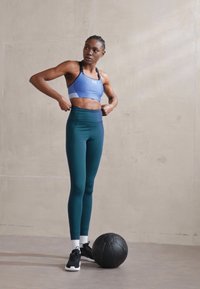 Sports bh i lyseblå med en hvid kant, parret med højtaljede teal leggings. Sorte sko og en sort træningsbold på gulvet.