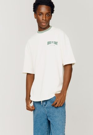 Cream-colored oversized t-shirt with green ribbed collar, featuring "HOUSE OF SUNNY" text in green and relaxed fit. Paired with blue denim pants.