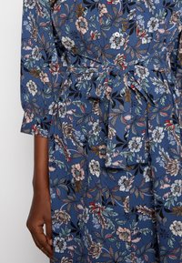 Květinové šaty z modré látky s páskem v oblasti pasu, dlouhými rukávy a knoflíkovými detaily. Vzor obsahuje růžové a bílé květy se zelenými listy.