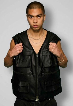 Uomo che indossa un gilet in pelle nero senza maniche con più tasche, che tiene i bordi del colletto, espressione neutra, su sfondo grigio semplice.