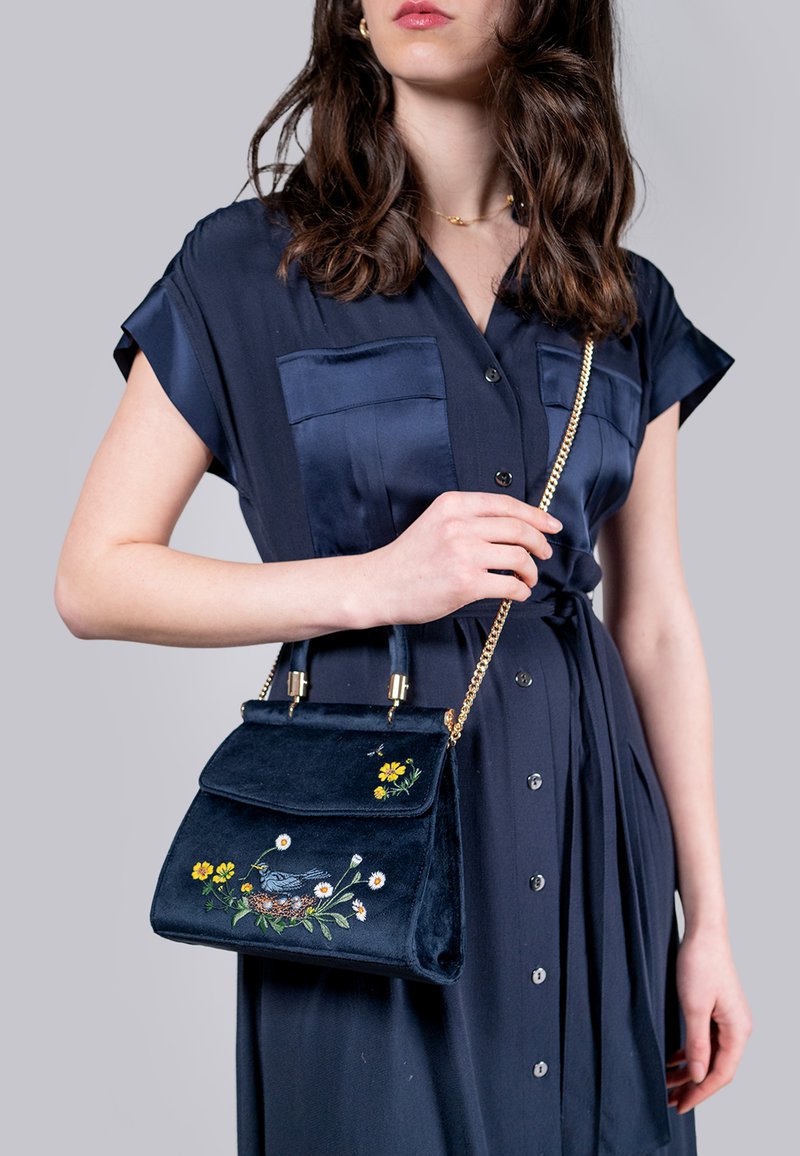 Bolso de terciopelo en navy con bordado floral, herrajes dorados, asas dobles y cadena para el hombro. Textura suave, forma estructurada.