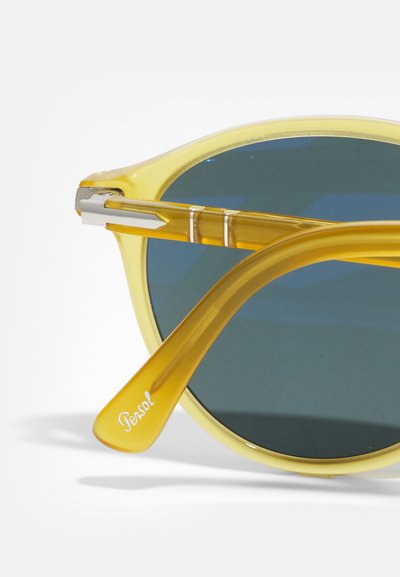Lunettes de soleil en plastique jaune avec des verres vert foncé. Présente des branches bicolores avec des accents en acier inoxydable et le logo "Persol" sur un côté.