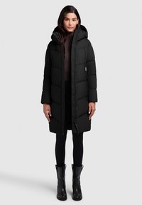 Schwarze Daunenjacke mit Kapuze, ausgestattet mit gesteppten Paneelen, einem Reißverschluss vorne und Druckknöpfen. Kombiniert mit schwarzen Leggings und Stiefeletten.