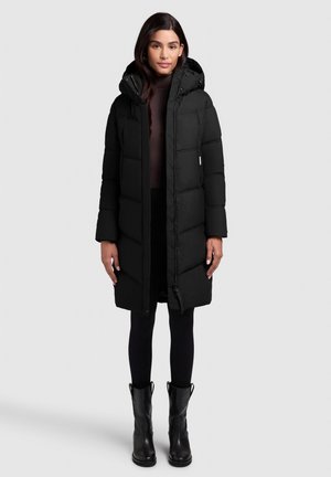 Zwarte pufferjas met capuchon, voorzien van gestikte panelen, een rits aan de voorkant en drukknoopsluiting. Gemaakt in combinatie met zwarte leggings en enkellaarsjes.