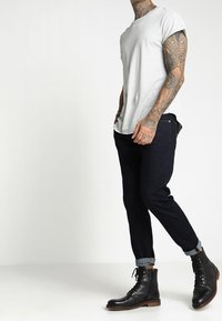 Muž v bílém tričku, tmavých zkrácených džínách, černých šněrovacích botách a s podrobnými tetováními na levém paži a krku.