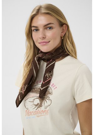 Junge Frau mit langem blondem Haar, die ein beiges Grafik-T-Shirt und einen braun gemusterten Schal trägt, der um ihren Hals gebunden ist.