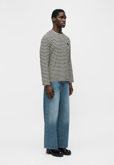 Άνδρας που στέκεται φορώντας μακρυμάνικο πουκάμισο με μαύρες και λευκές ρίγες, φαρδιά μπλε τζιν και μαύρα παπούτσια με τακούνι σε απλό φόντο.