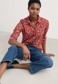 Giovane donna seduta a terra con una camicia rossa a fiori, jeans blu a vita alta, cintura nera e scarpe nere su sfondo semplice.