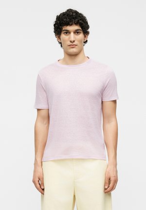 Mand med krøllet mørkt hår iført lyserød kortærmet t-shirt og cremefarvede bukser mod en ensfarvet hvid baggrund.