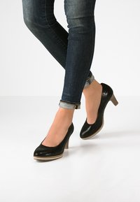 Svarta lackläderklackar med rund tå, låg klack och en ljusfärgad plattformdetalj, bärs med mörkblå uppvikta jeans.