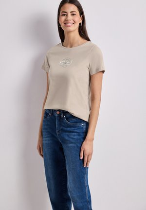 Kvinde med langt mørkt hår iført beige kortærmet skjorte med grafik og blå jeans, står mod en ensfarvet lys baggrund og smiler.