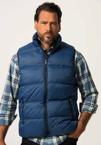 Tummansininen puffatusti takki, jossa on korkea kaulus, vetoketjusuljenta ja kaksi sivutaskua; päällä ruudullinen nappikauluspaita, jossa on vihreitä ja sinisiä yksityiskohtia.