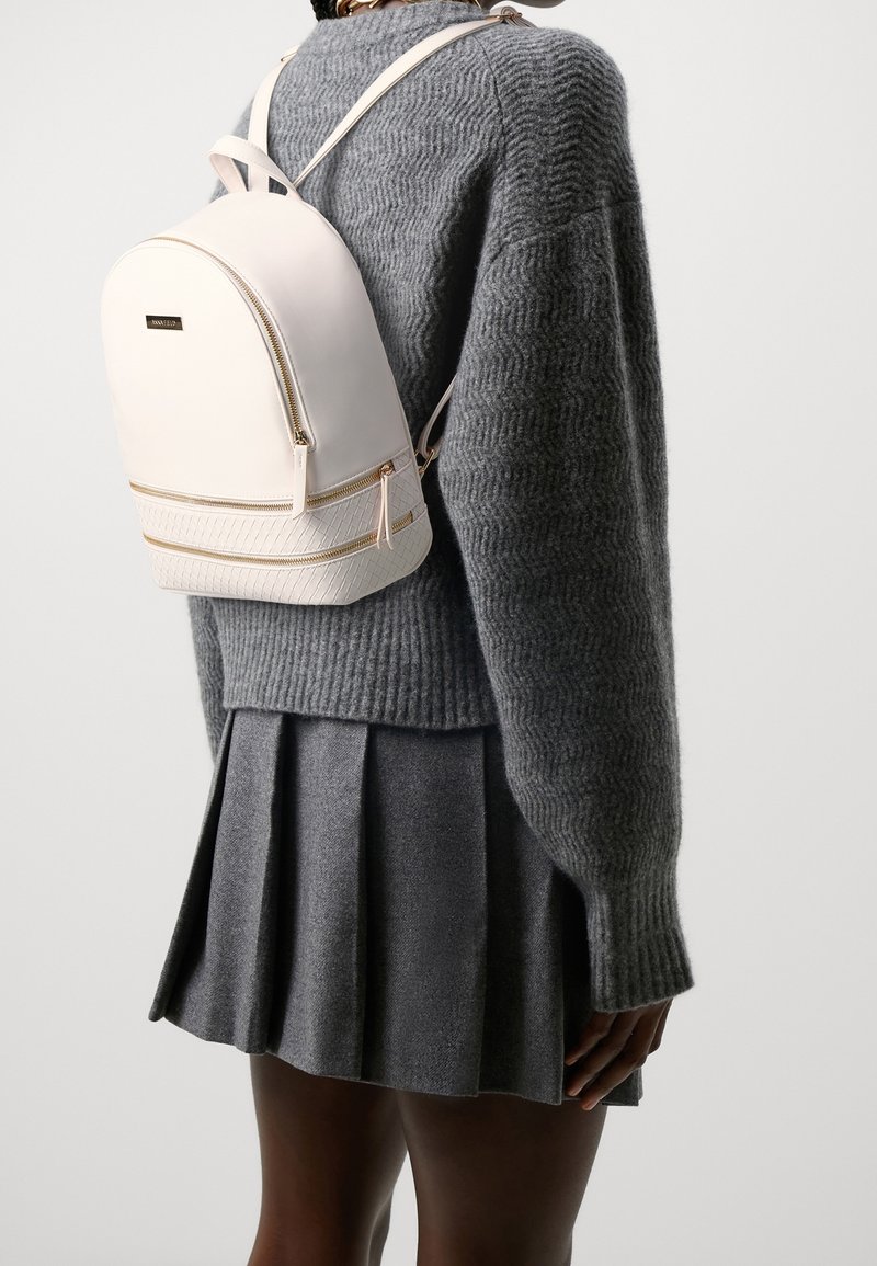 Mochila rosa claro con un acabado suave, cremalleras doradas y un panel inferior texturizado. Usada sobre un suéter de punto gris y una falda plisada.