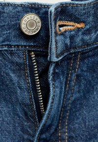 Gros plan d'un jean denim bleu montrant une fermeture éclair partiellement ouverte, un bouton en métal avec texte et une couture orange visible autour de la ceinture.
