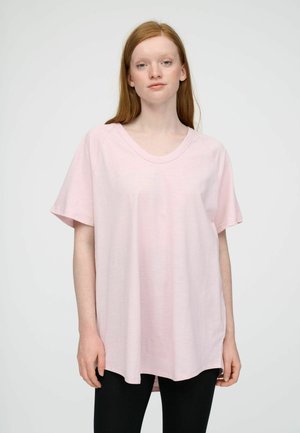 Jonge vrouw met lang rood haar die een los lichtroze T-shirt en zwarte broek draagt tegen een effen witte achtergrond.