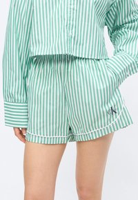 Zaļas un baltas vertikālas svītras šorti ar elastīgu vidukli un sānu kabatām, valkāti kopā ar atbilstošu garroku pogājamu kreklu.