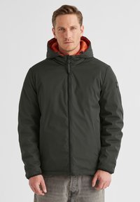 Dunkelgrüne Zip-Jacke mit Kapuze, ausgestattet mit einem orangefarbenen Innenfutter, glatter Textur und einem Logopatch am linken Ärmel. Mit Fronttaschen.