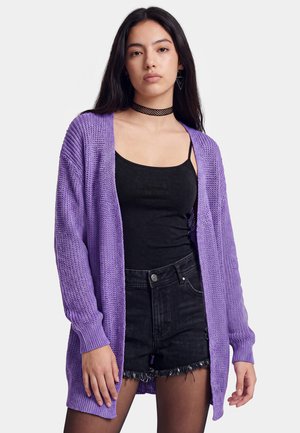 Donna con un cardigan di lana viola sopra una canotta nera, shorts di jeans neri a vita alta, calze nere, choker nero e orecchini geometrici.