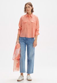 Koraalkleurig overhemd met knopen en twee borstzakken, gecombineerd met lichtblauwe recht gesneden jeans. Het model houdt een lichtgewicht roze sjaal met patroon vast.