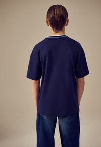 Meriinse sinine puuvillane T-särk, millel on ümarsärk, lühikesed varrukad ja valge topelttriip kaelusel, kombineeritud siniste teksadega.