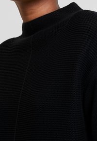 Pull noir en tricot avec un col haut, présentant un motif texturé côtelé et des lignes épurées. Le tissu semble doux et extensible.