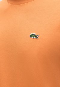 Πορτοκαλί ύφασμα με μικρό πράσινο κεντητό λογότυπο κροκοδείλου με κόκκινες λεπτομέρειες στην πάνω περιοχή του στήθους.